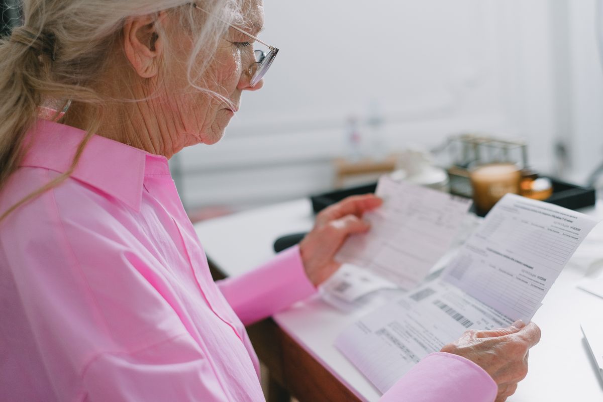 An older woman with silver hair and glasses reads a benefits statement at her table, the kind of letter that brings the 2027 Social Security cost-of-living increase.