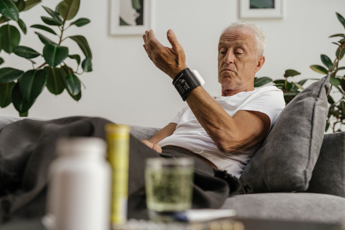 A white-haired man sits on a gray couch at home, reading a wrist blood pressure monitor on his raised arm, with a pill bottle and a glass of water in the foreground, the daily routine a Utah retiree with heart disease or diabetes manages under a Medicare Chronic Condition Special Needs Plan.