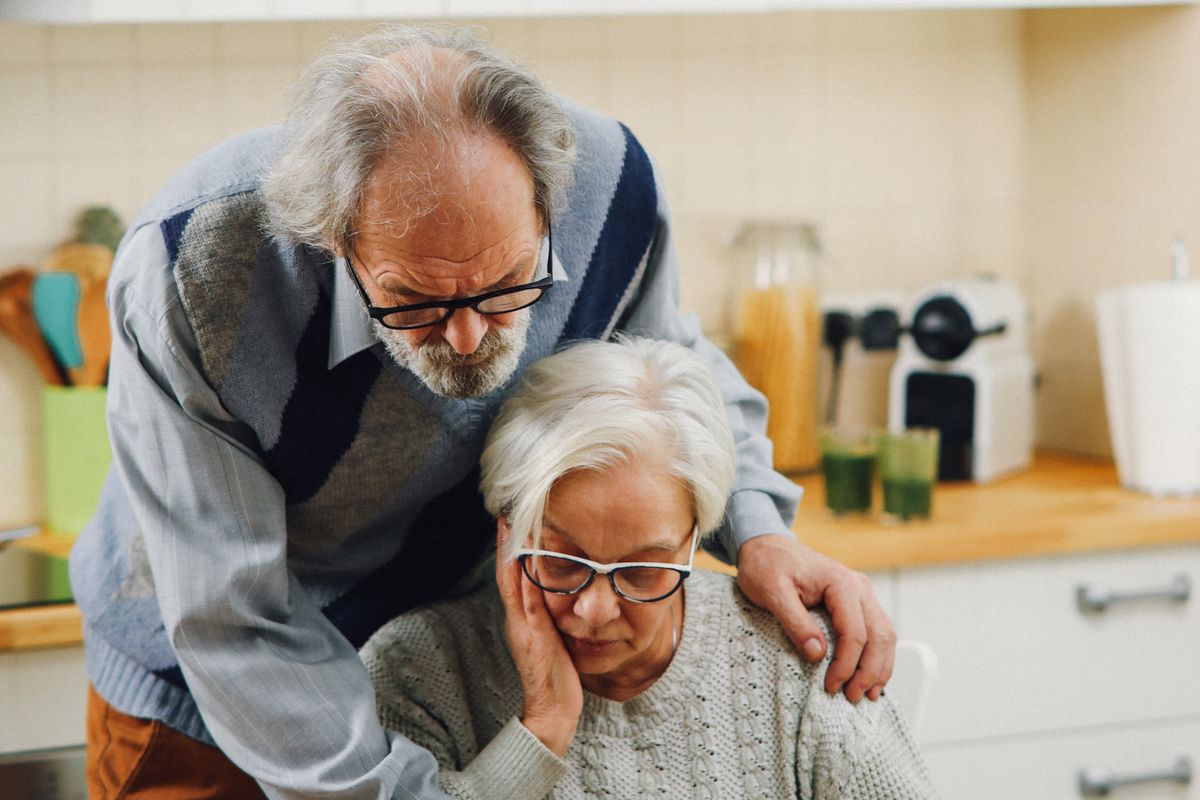 A gray-haired man leans over his wife's shoulder at their kitchen table as they look down at household paperwork, working out a retirement budget.