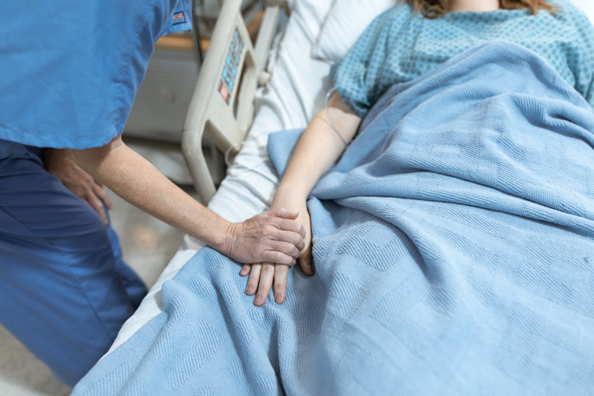 A hospital staff member in scrubs holding the hand of a patient resting in a hospital bed, illustrating a stay where inpatient versus observation status is unclear.