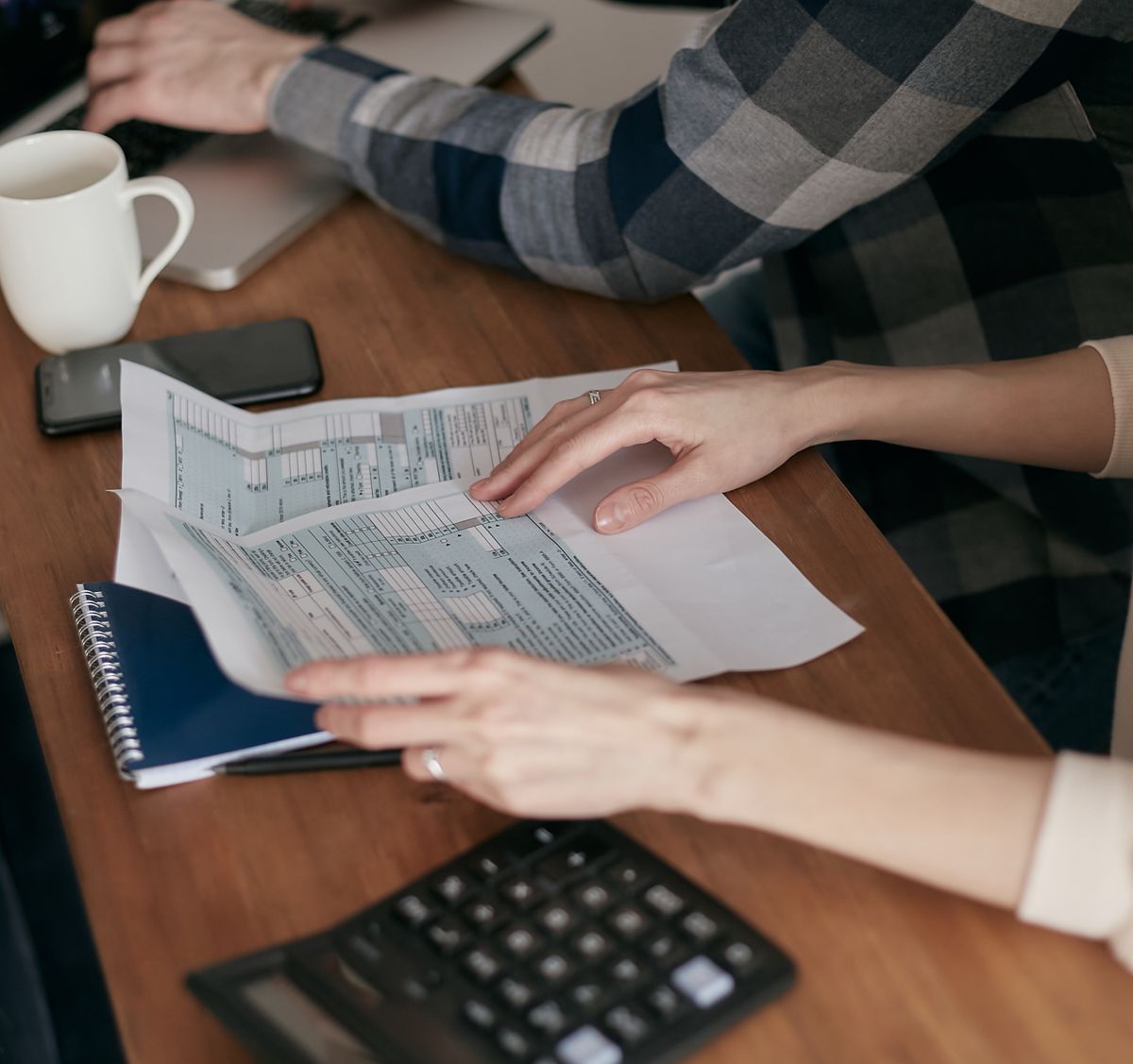 Hands hold printed federal tax forms on a wooden table beside a calculator, notebook and coffee mug, working out how annuity income is taxed.