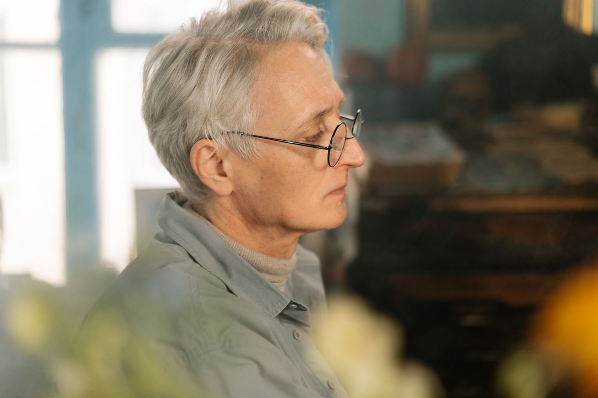 An older man with gray hair and round wire-rimmed glasses sits in profile by a bright window, the age at which cataracts cloud the lens and Medicare Part B pays for the surgery that replaces it, a common procedure for Utah retirees.