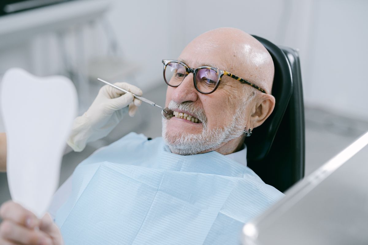 An older adult in a dental chair reviewing his teeth in a hand mirror — routine dental care that Original Medicare does not cover for Utah retirees.