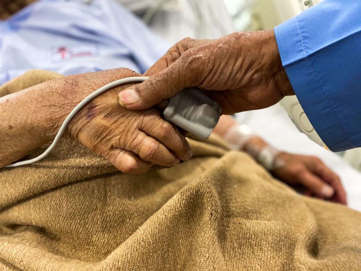 An older patient's hand rests on a tan blanket with a fingertip monitor clipped on while a visitor in a blue shirt holds it, the kind of hours-long treatment session a Utah family plans around when Medicare covers dialysis for kidney failure.