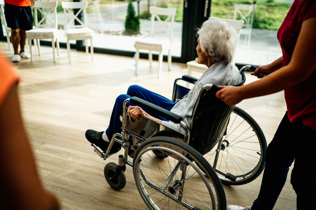 A caregiver pushes a white-haired woman in a manual wheelchair across a sunlit room, the kind of durable medical equipment Medicare Part B rents for 13 months before ownership passes to the Utah retiree using it.