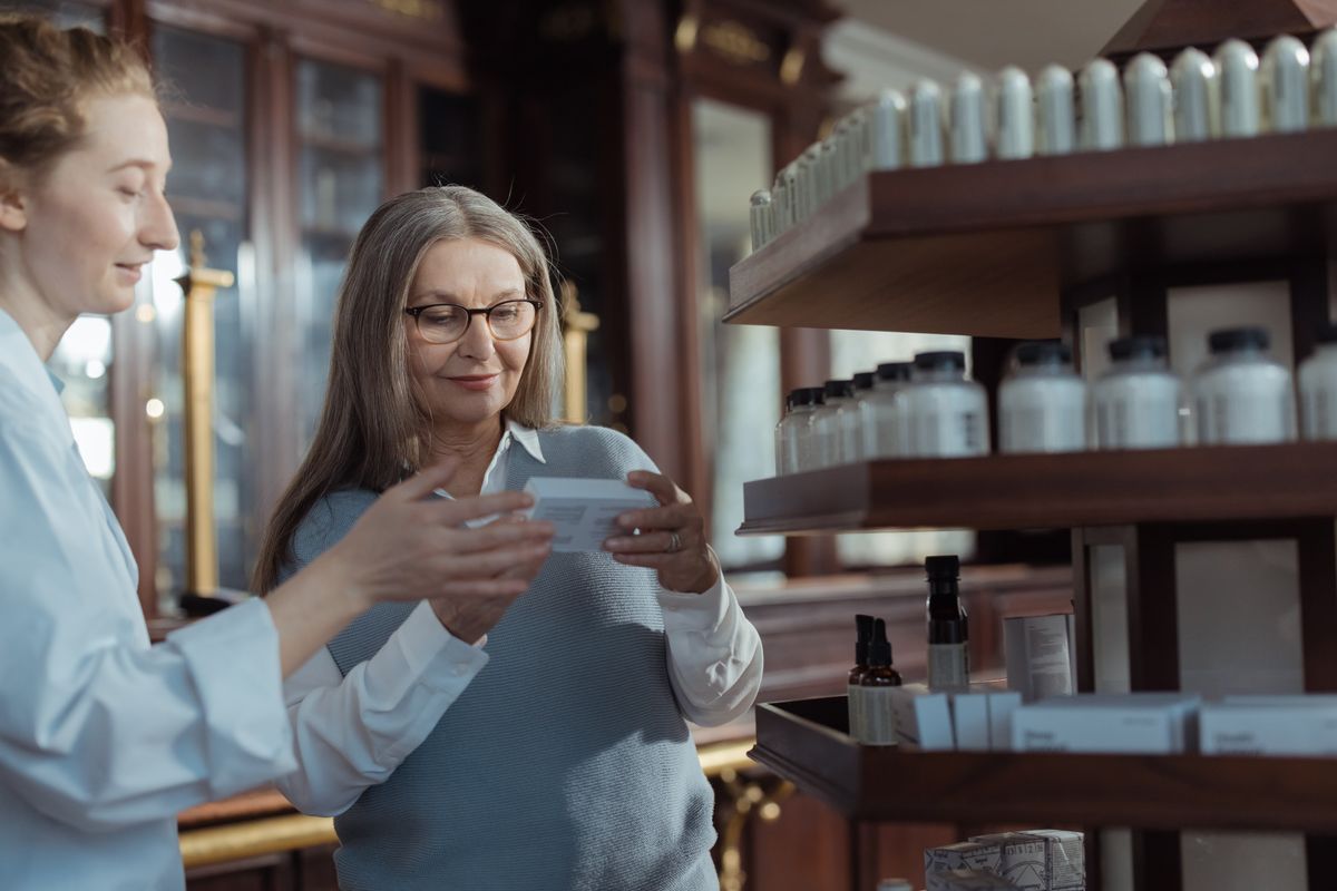 An older woman with glasses and long gray hair reads the label on a medication box while a pharmacist in a white coat explains it, the kind of pharmacy conversation a Utah retiree has when a GLP-1 drug is filled through Medicare.