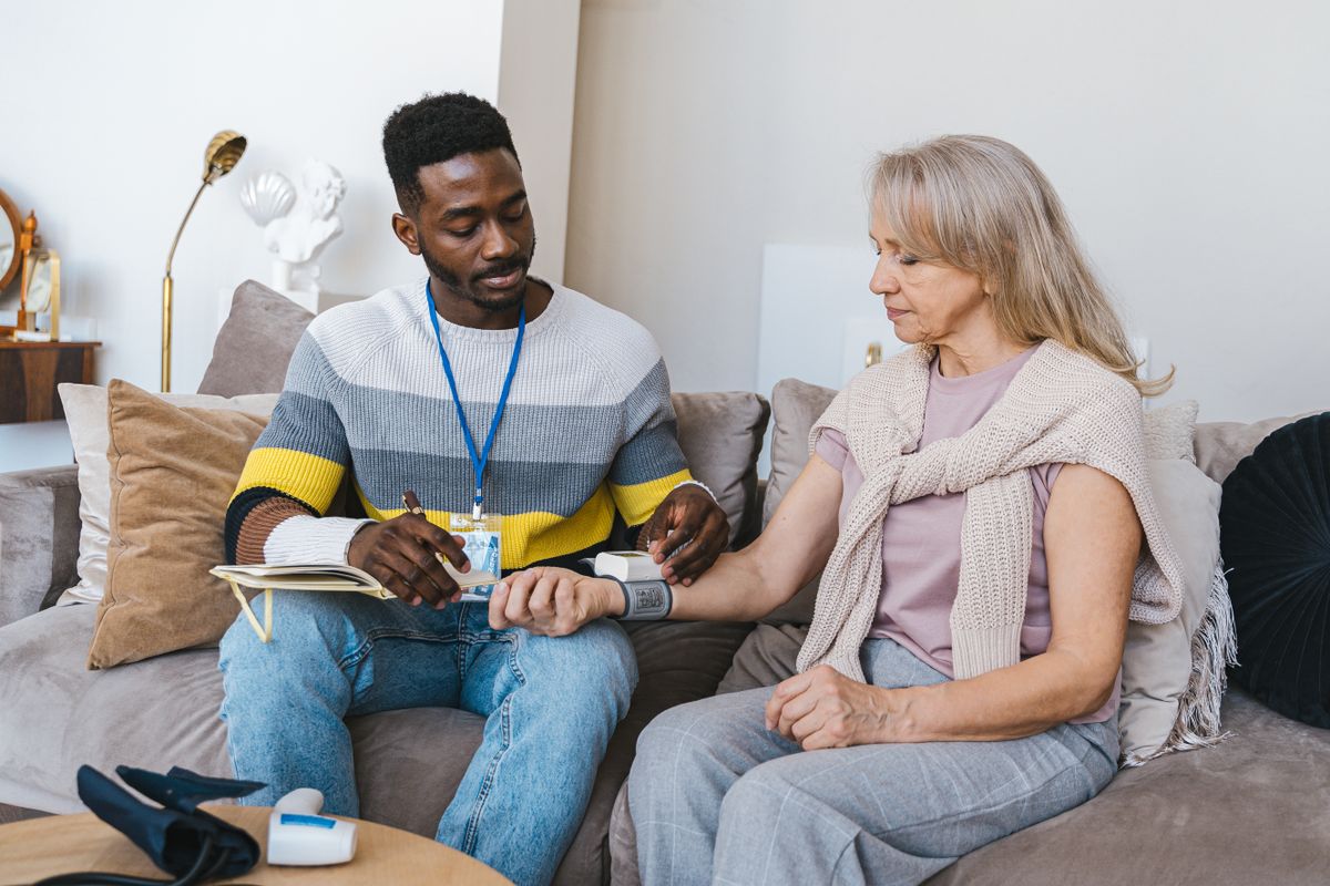 A home health nurse with a lanyard sits beside an older woman on her living-room couch, wrapping a blood pressure cuff on her wrist and writing in a notebook, the kind of skilled nursing visit Medicare covers at home in Utah.