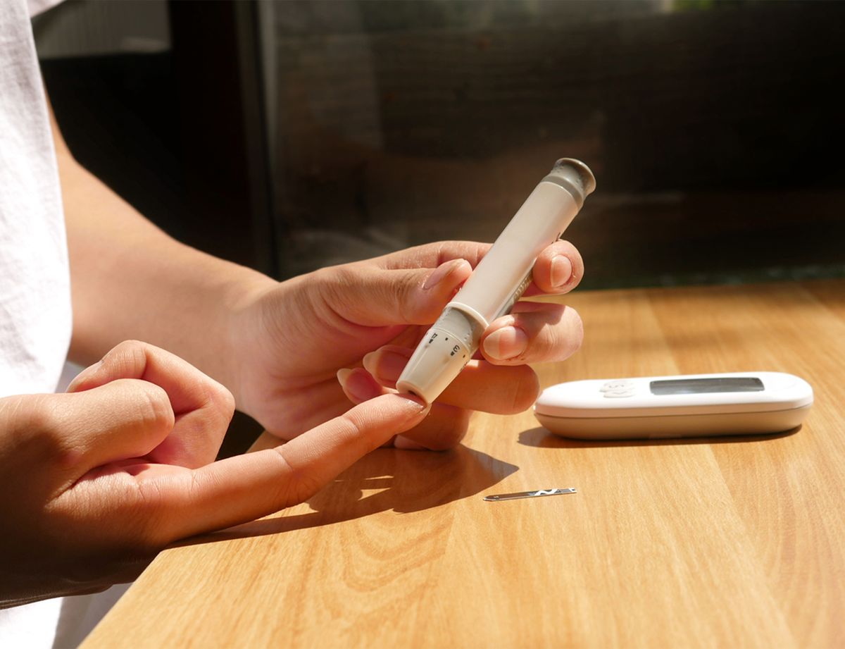 A Utah retiree's hands use a lancing device to check blood sugar at a sunlit wooden kitchen table, with a glucose meter and test strip beside them.