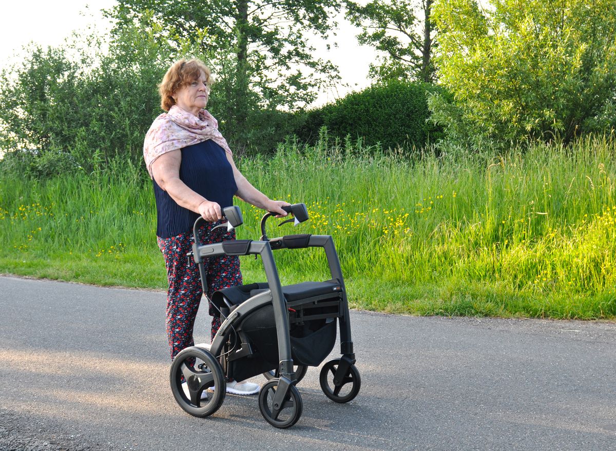 An older woman walks along a quiet country road behind a four-wheeled rollator walker on a summer evening, the kind of recovery walk that follows a Medicare-covered knee or hip replacement for a Utah retiree.
