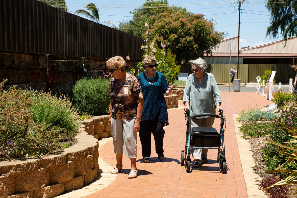 Three retired women in their seventies walk and talk together along a sunny garden path, one pushing a rollator, the kind of friends who remind each other that a Medicare-covered screening mammogram is due every year for Utah women.