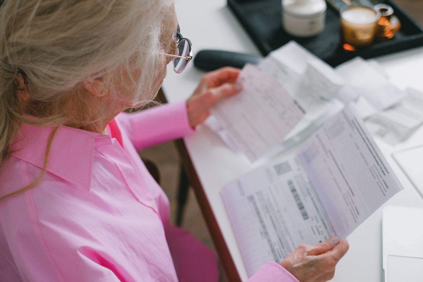 An older Utah woman in reading glasses looks through a stack of insurance statements and a plan notice at her desk, with prescription containers beside her.