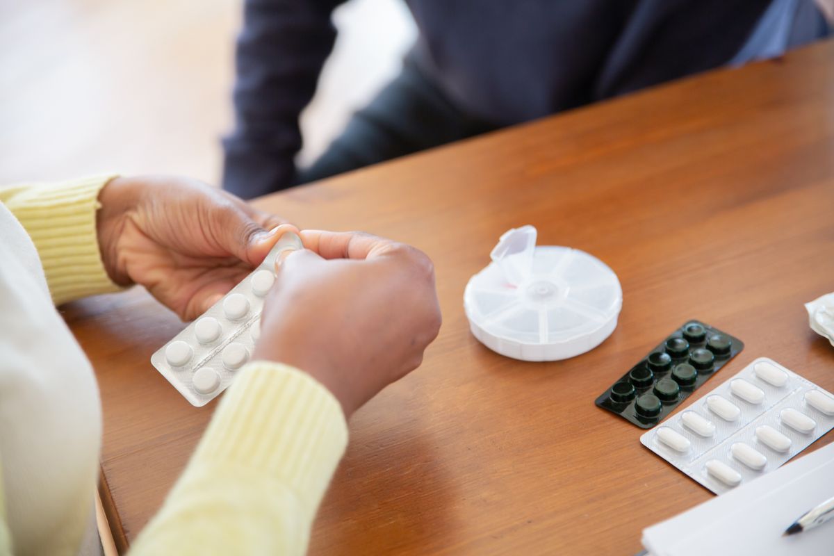 Close view of a person's hands pressing tablets out of a blister pack beside a round weekly pill organizer on a wooden table, the routine a Utah retiree's Medicare Part D drug plan pays for.