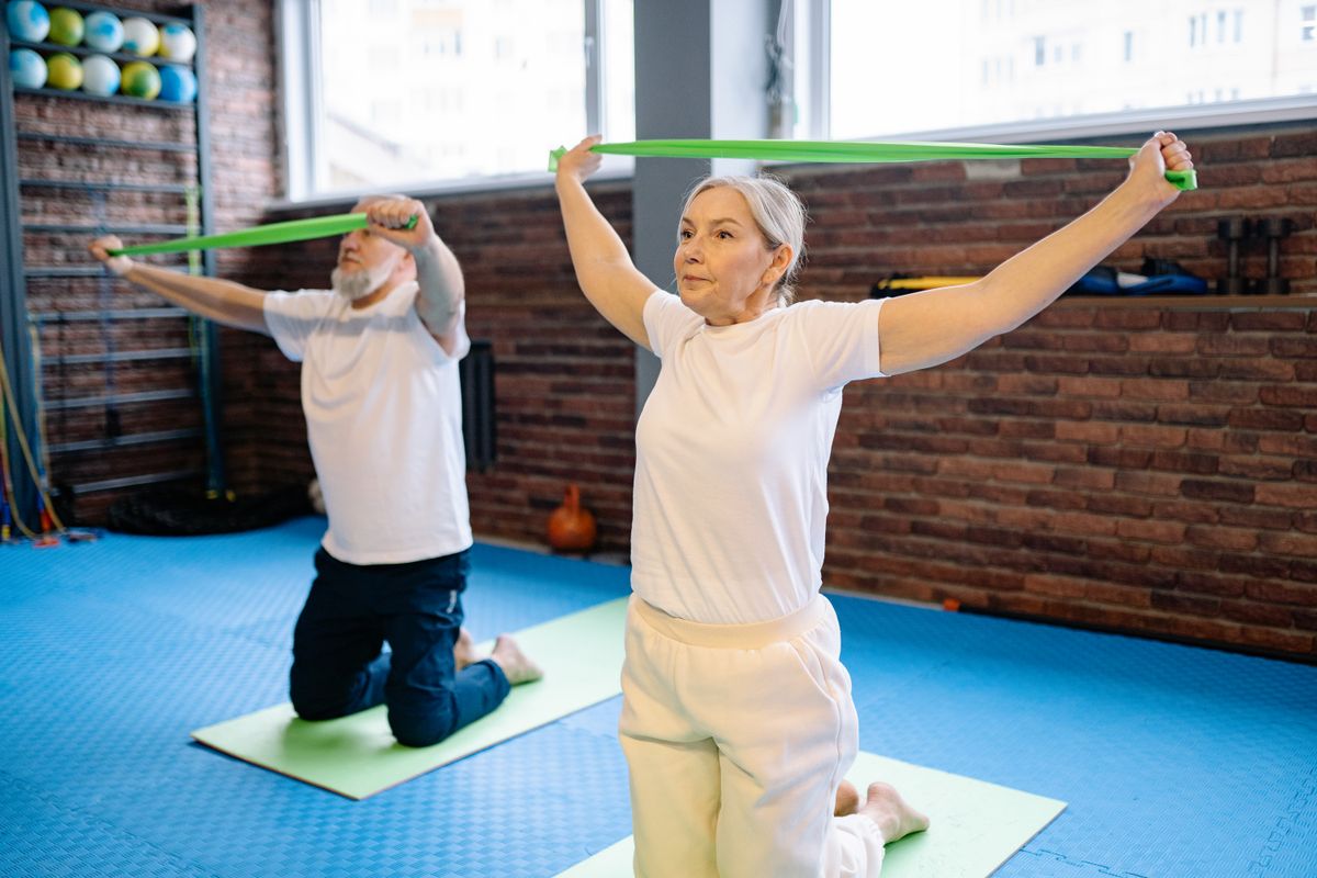 A woman and a man in their sixties kneel on exercise mats in a bright brick-walled gym, each stretching a green resistance band overhead, the kind of supervised strength and balance work a Utah retiree gets in Medicare-covered outpatient physical therapy.