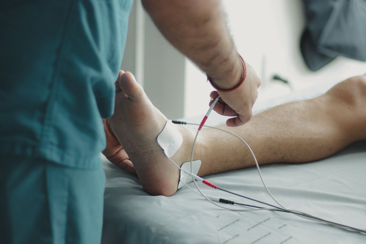 A clinician in teal scrubs steadies a patient's bare foot on an exam table while attaching small sensor pads near the ankle, the kind of foot and nerve check that decides whether Medicare Part B treats a Utah retiree's foot care as covered treatment or as routine care.