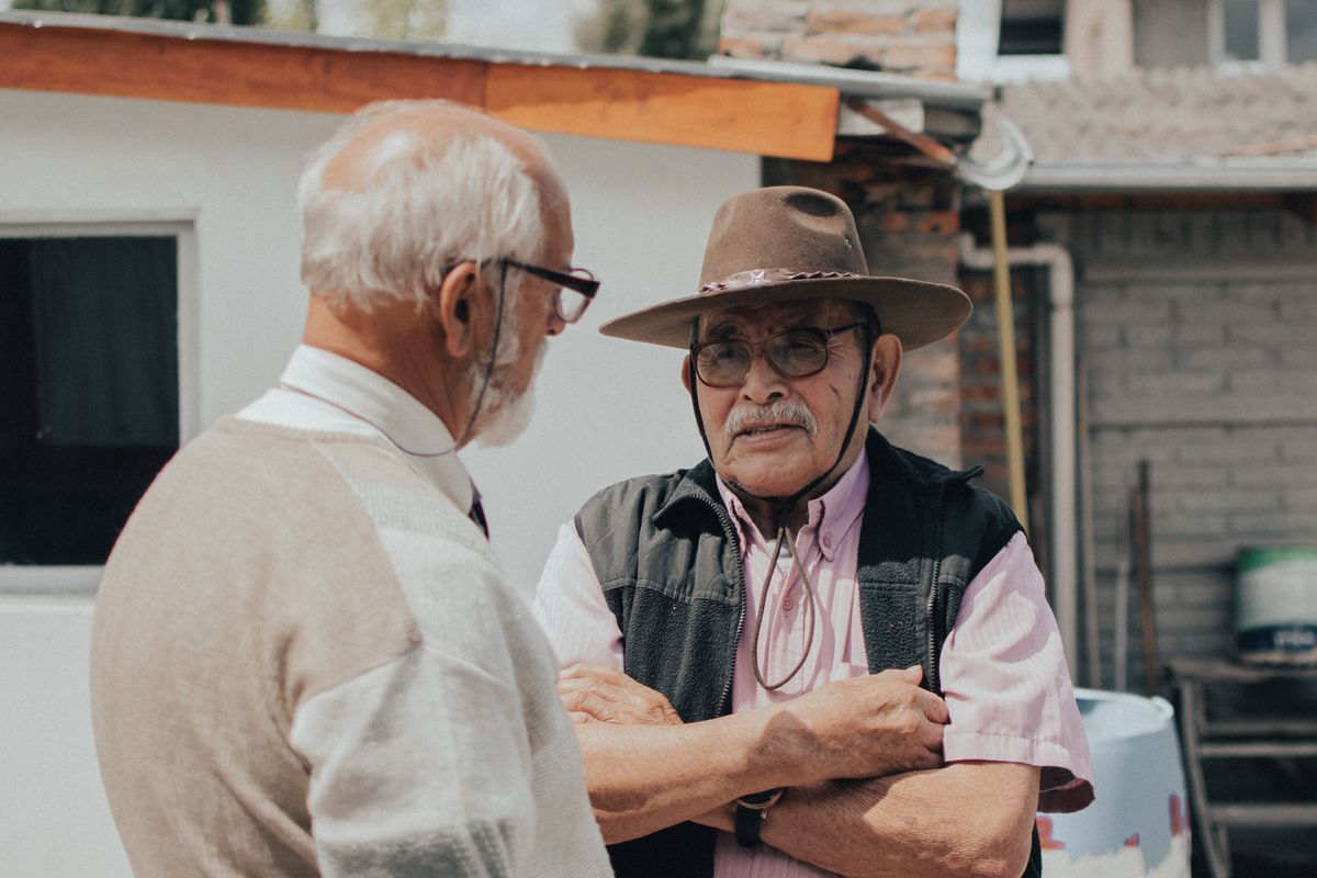 Two retired men in their seventies, one in a western hat and vest with his arms folded, talk in the sun outside a small-town building, the kind of conversation in which a Utah man learns his friend's Medicare PSA test was free and the biopsy was not.