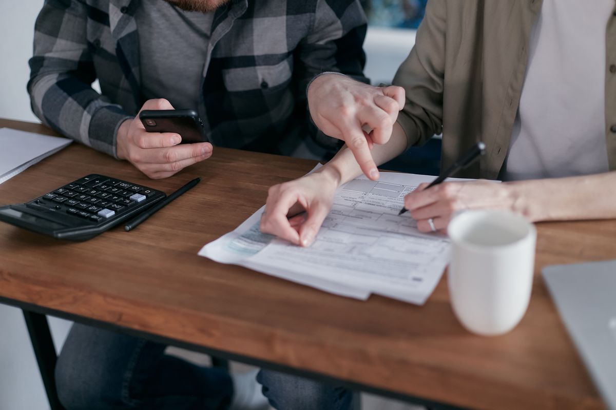 Two people work through printed federal tax forms at a wooden table, one pointing at a line, with a calculator, pen and coffee mug nearby, figuring out the senior deduction.