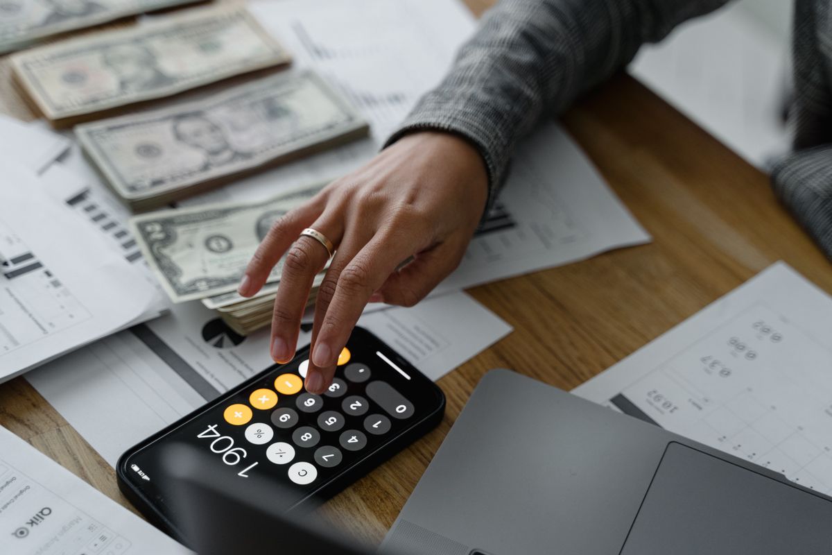 A hand taps a phone calculator beside stacks of U.S. dollar bills and printed statements on a wooden table, working out where to keep retirement cash.