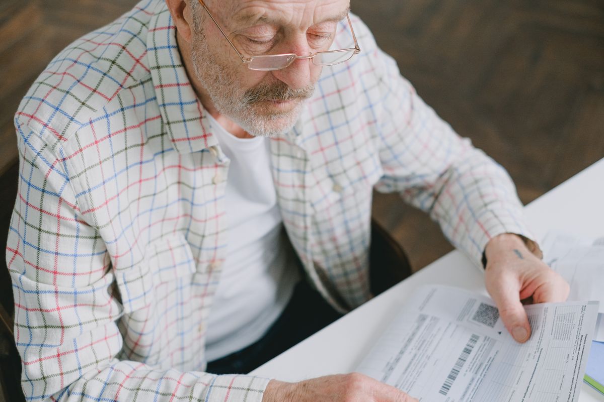 A retired man with a gray beard and glasses reads through tax paperwork at a table, reviewing year-end tax moves before December 31.